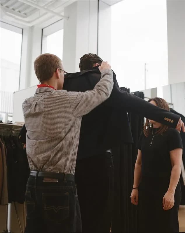 Two people assist a man in trying on a tailored jacket in a studio setting, highlighting fit, structure, and garment adjustment during the styling process.