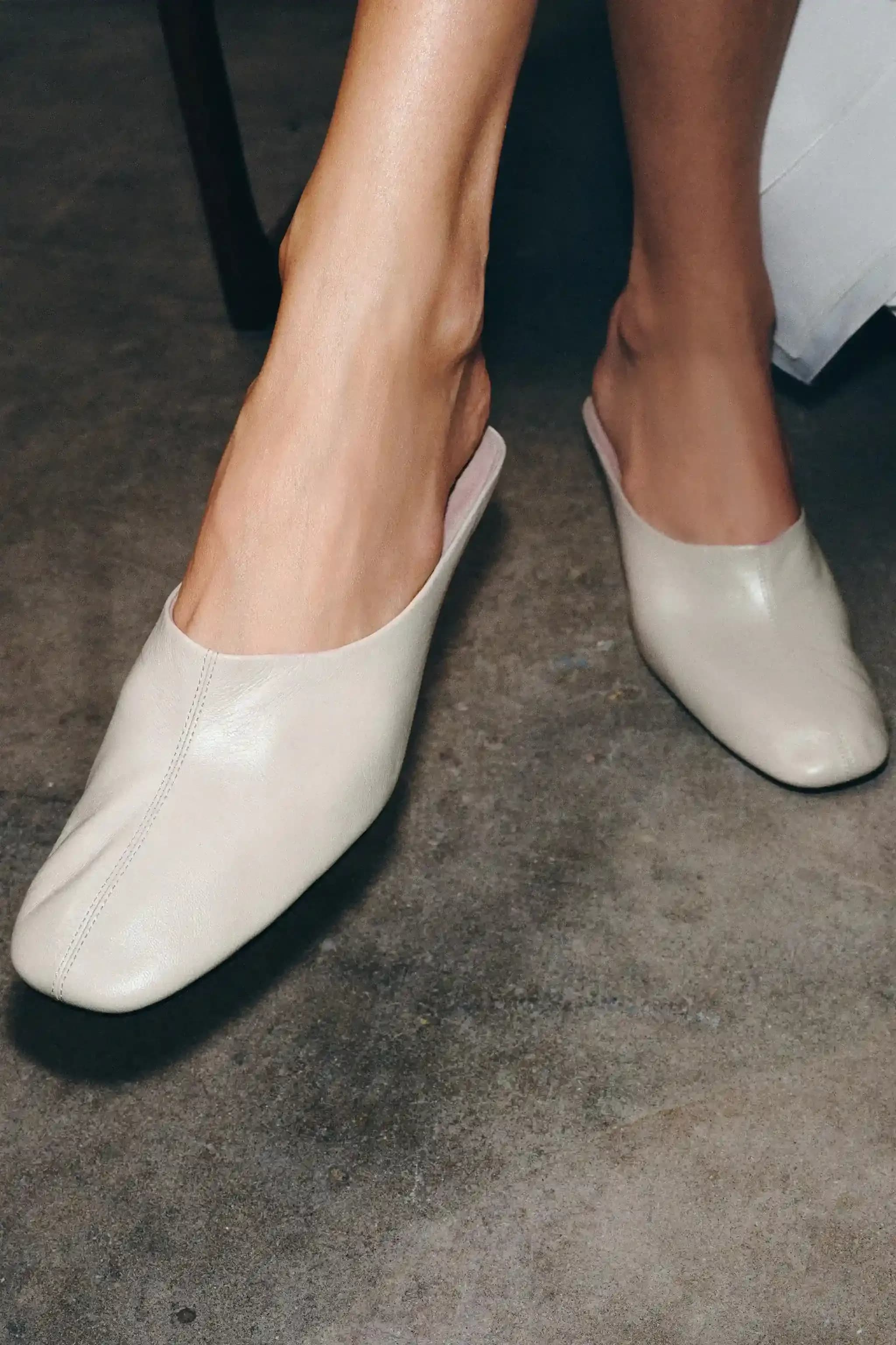 Close-up of feet wearing minimalist cream leather slip-on shoes, highlighting clean design and soft texture against a concrete floor.