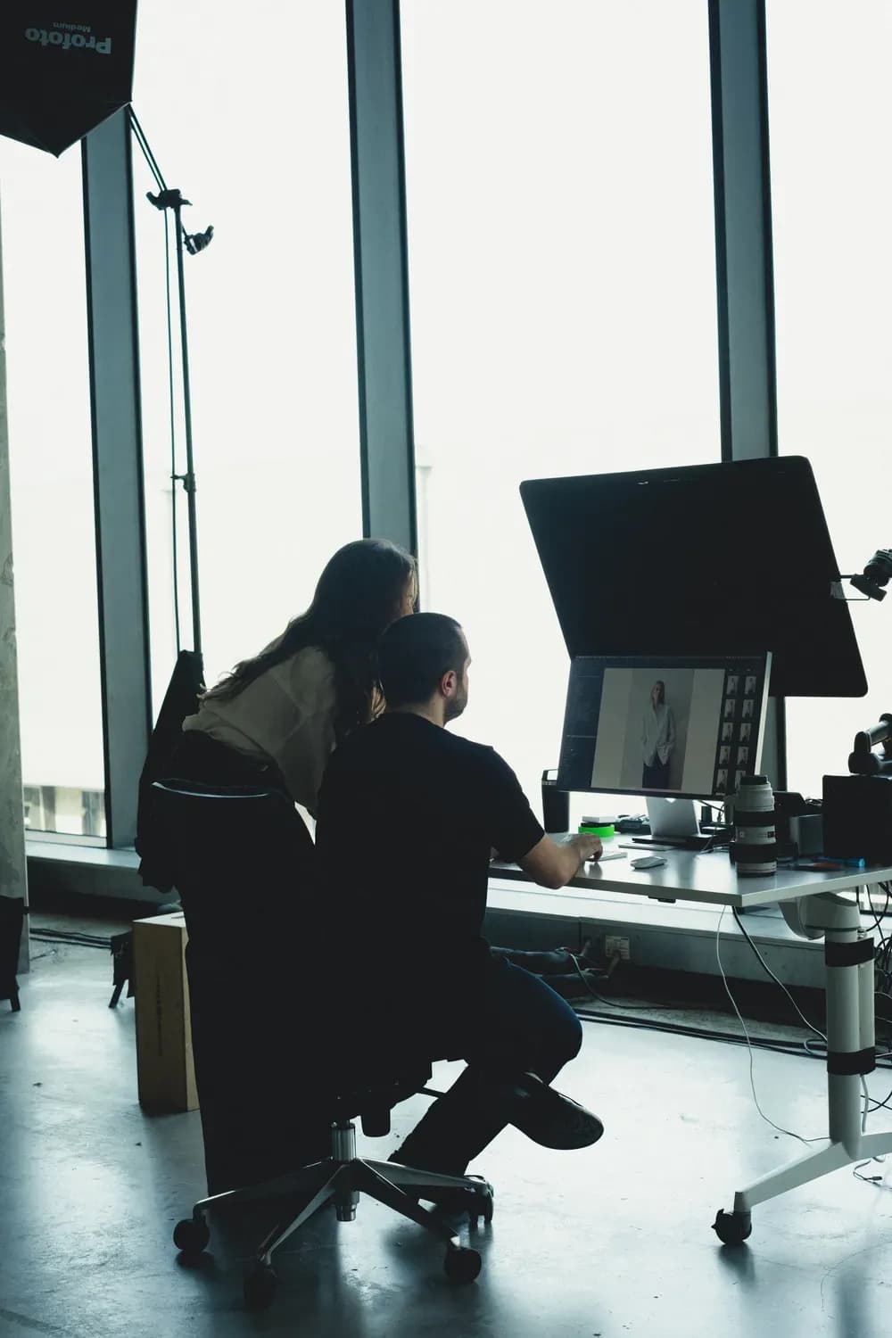 Deux personnes dans un studio, examinant des images de mode sur un ordinateur, entourées de matériel d'éclairage photographique et de grandes fenêtres.