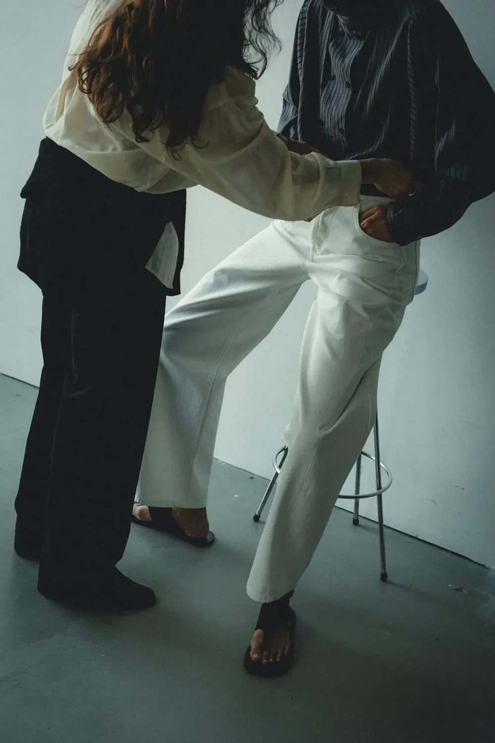 A stylist adjusts the fit of light trousers on a seated model, highlighting tailoring, drape, and garment construction in a studio setting.