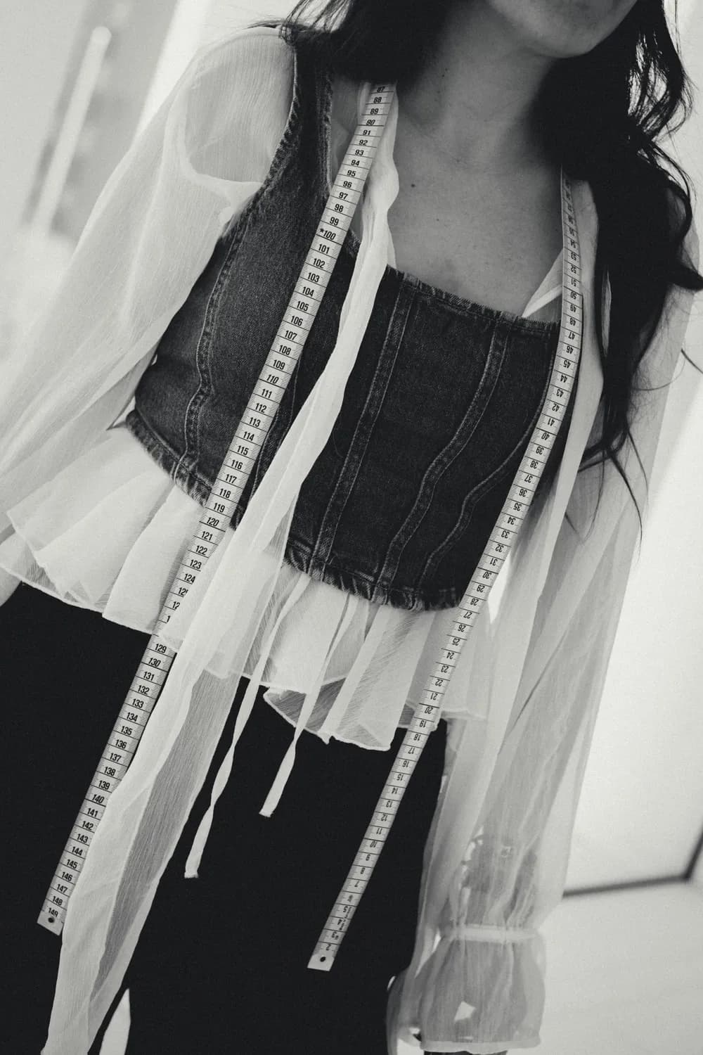 Black-and-white close-up of a person wearing a layered outfit with a denim corset and sheer blouse, with a measuring tape draped around their neck.