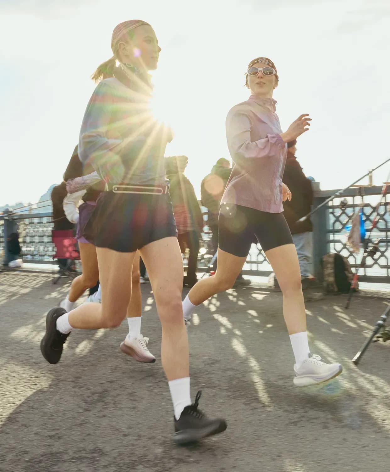 Deux femmes font leur jogging en extérieur, vêtues de tenues sportives coordonnées — vestes légères, shorts ajustés, chaussettes blanches et chaussures de course — le long d'un chemin ensoleillé au bord de la rivière, avec d'autres coureurs et des éléments de la ville en arrière-plan.