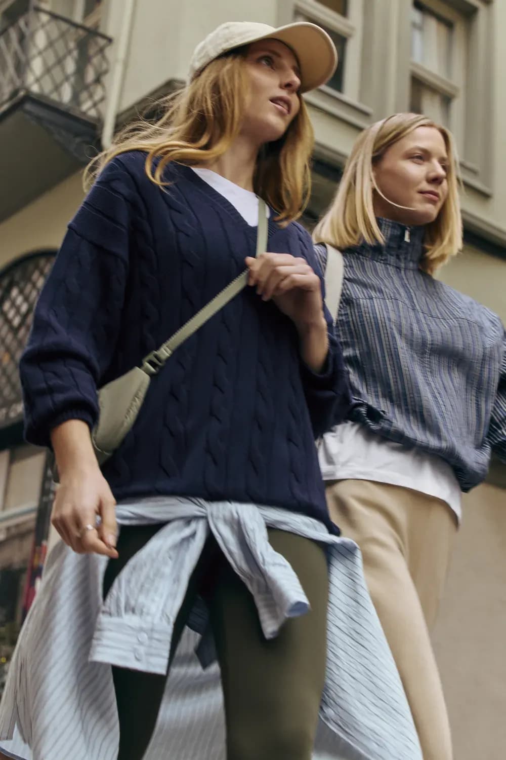 Deux femmes marchent à l'extérieur, vêtues de tenues décontractées superposées : l'une porte un pull en maille bleu marine, une casquette, un legging et une chemise nouée à la taille, l'autre un haut rayé à col montant et un pantalon clair ; elles transportent de petits sacs dans un cadre urbain.