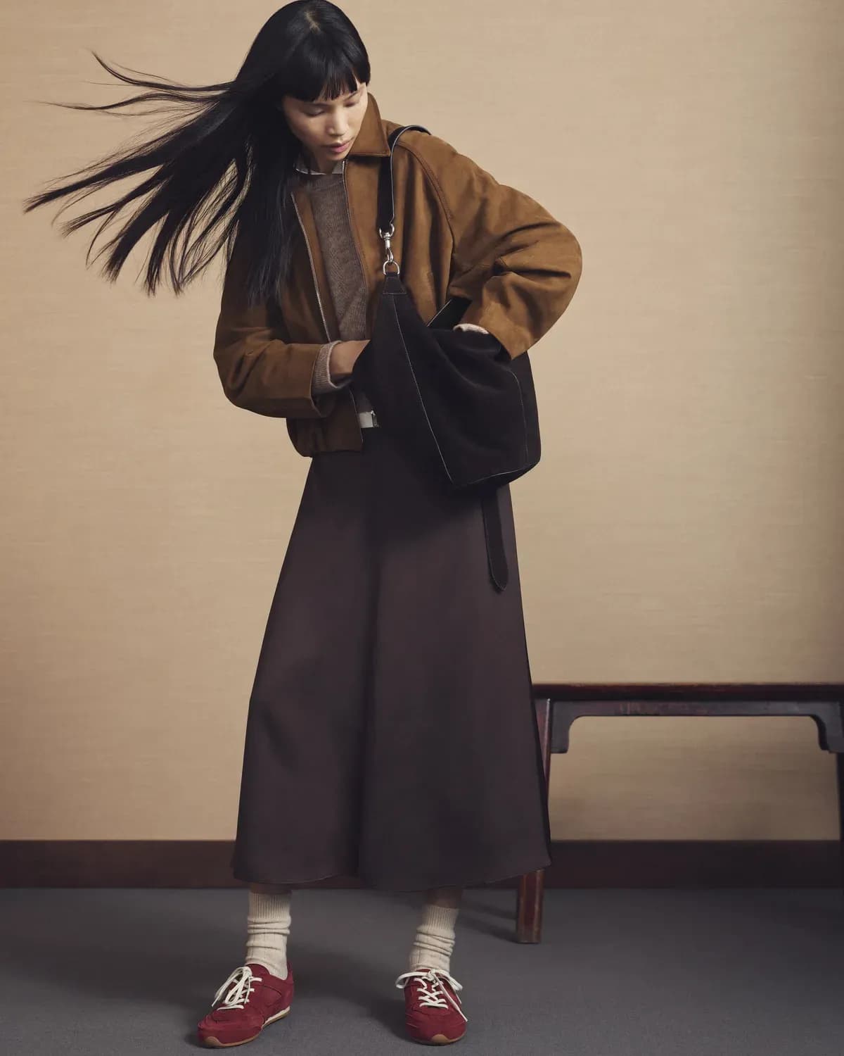 Une femme vêtue d'une veste marron, d'une jupe longue et de baskets rouges regarde dans un sac à bandoulière noir, ses cheveux bougeant tandis qu'elle se tient debout dans un décor intérieur minimaliste.