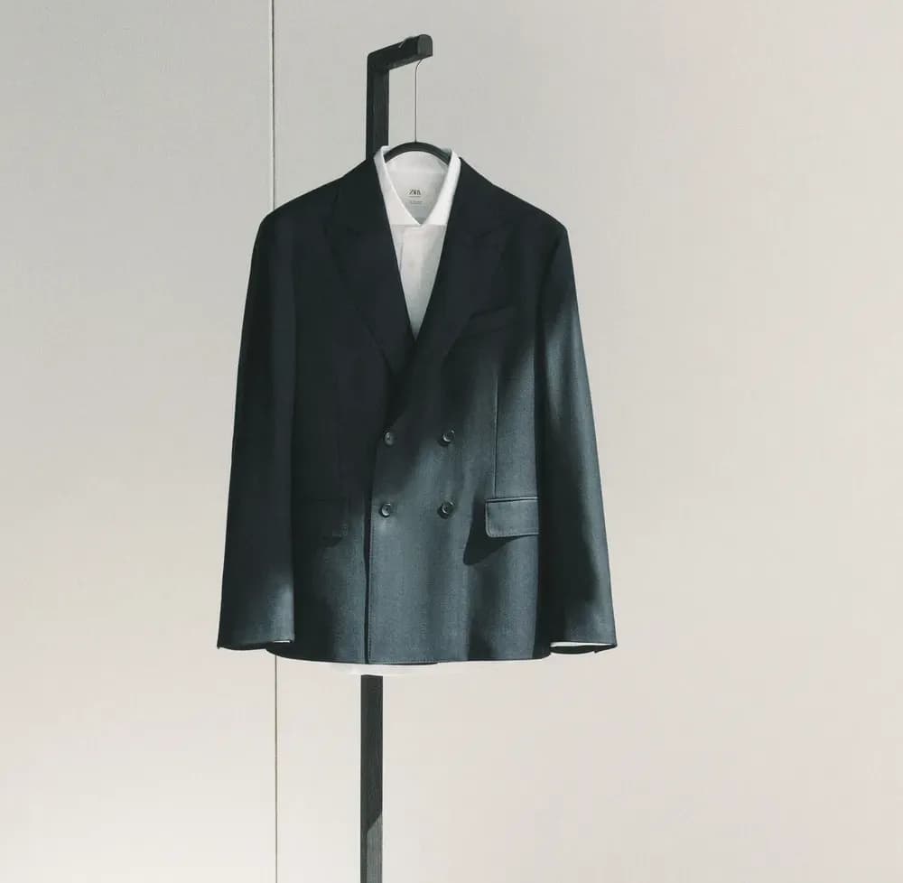 Un blazer croisé foncé structuré, porté sur une chemise blanche impeccable, est présenté sur un présentoir minimaliste, mettant en valeur la coupe, la symétrie et la texture raffinée du tissu dans un cadre épuré et moderne.