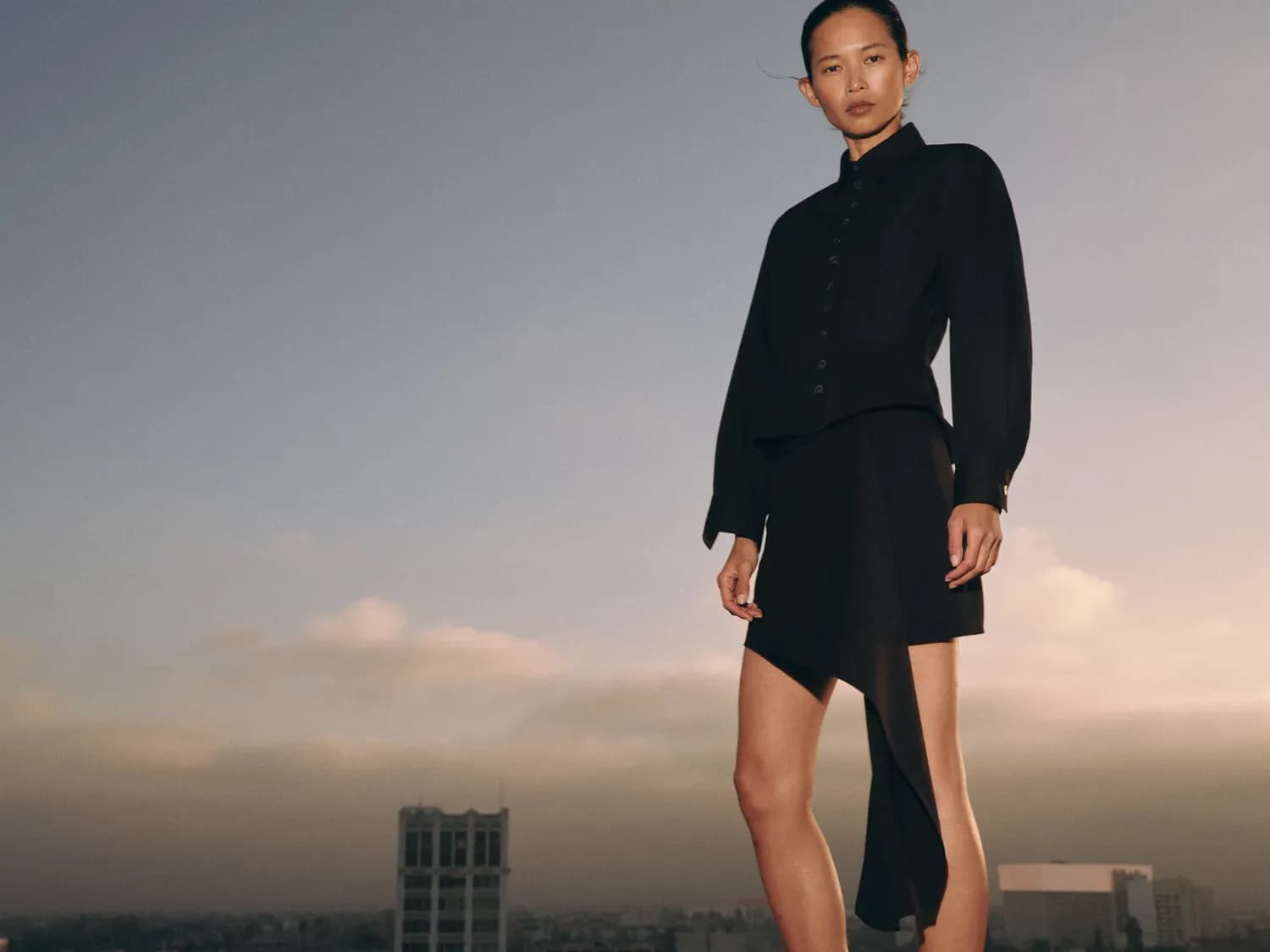 A woman wears a structured black outfit featuring a fitted button-up top and an asymmetrical skirt, highlighting sharp tailoring, fluid draping, and a sleek, modern silhouette against an open sky backdrop.