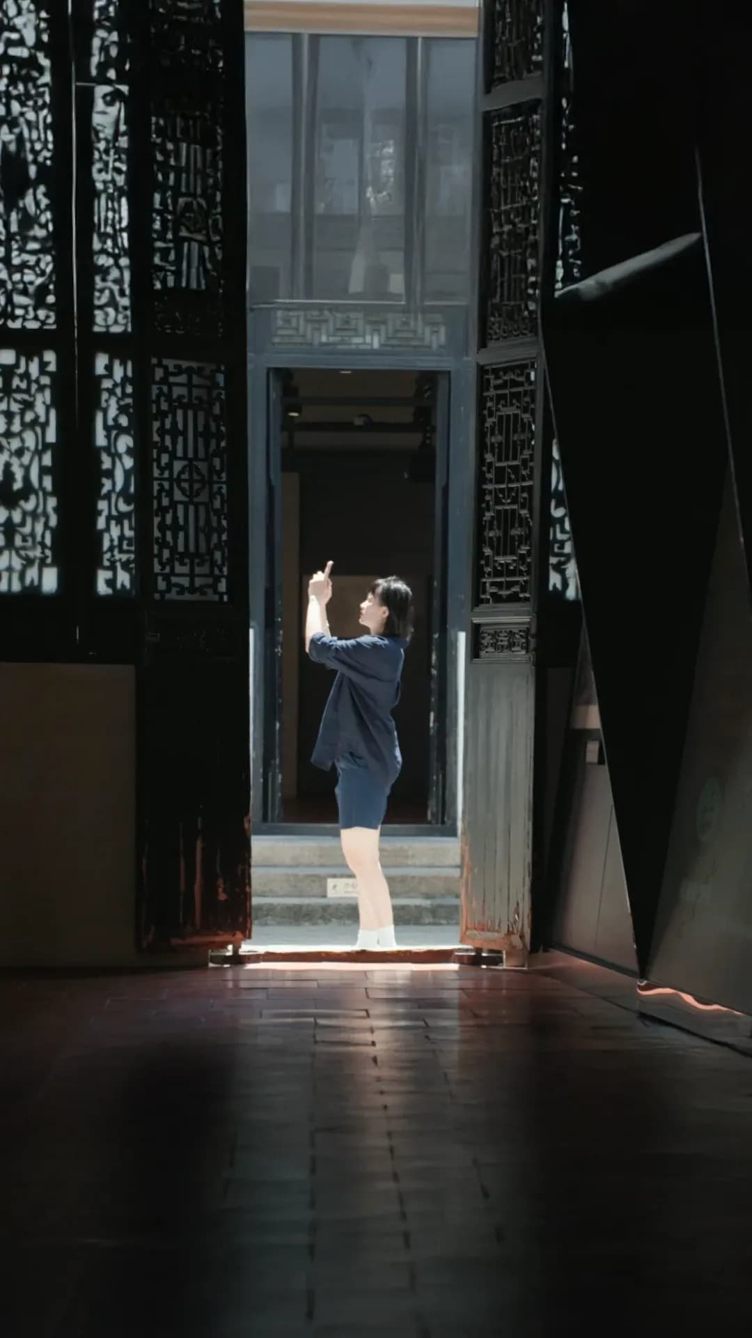A person stands in a doorway wearing a relaxed dark outfit, highlighted by natural light, emphasizing simple silhouettes and fluid movement within an architectural setting.