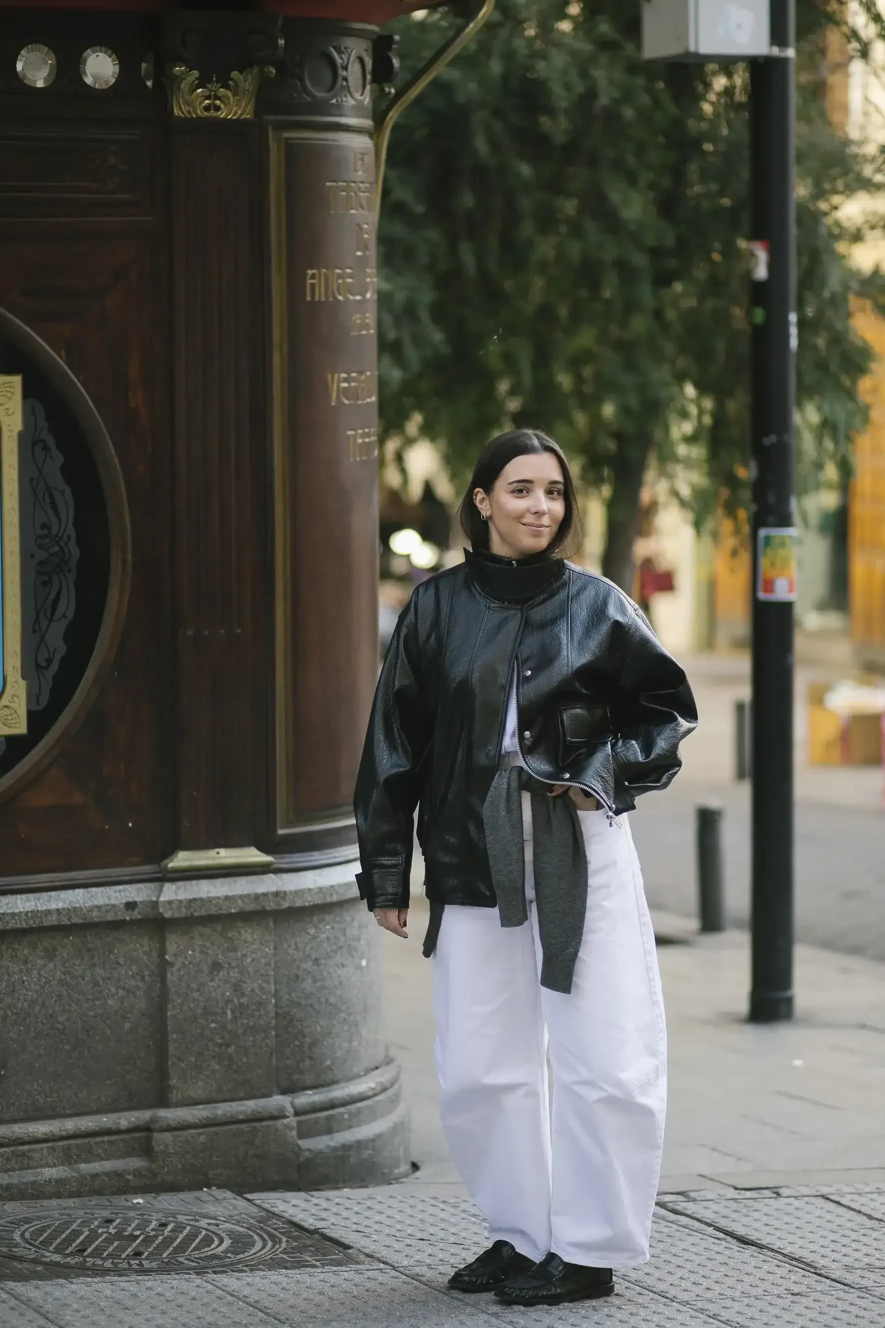 A woman stands on a city street wearing a black leather jacket paired with wide white trousers, creating a contrast between structured outerwear and relaxed, voluminous silhouettes.