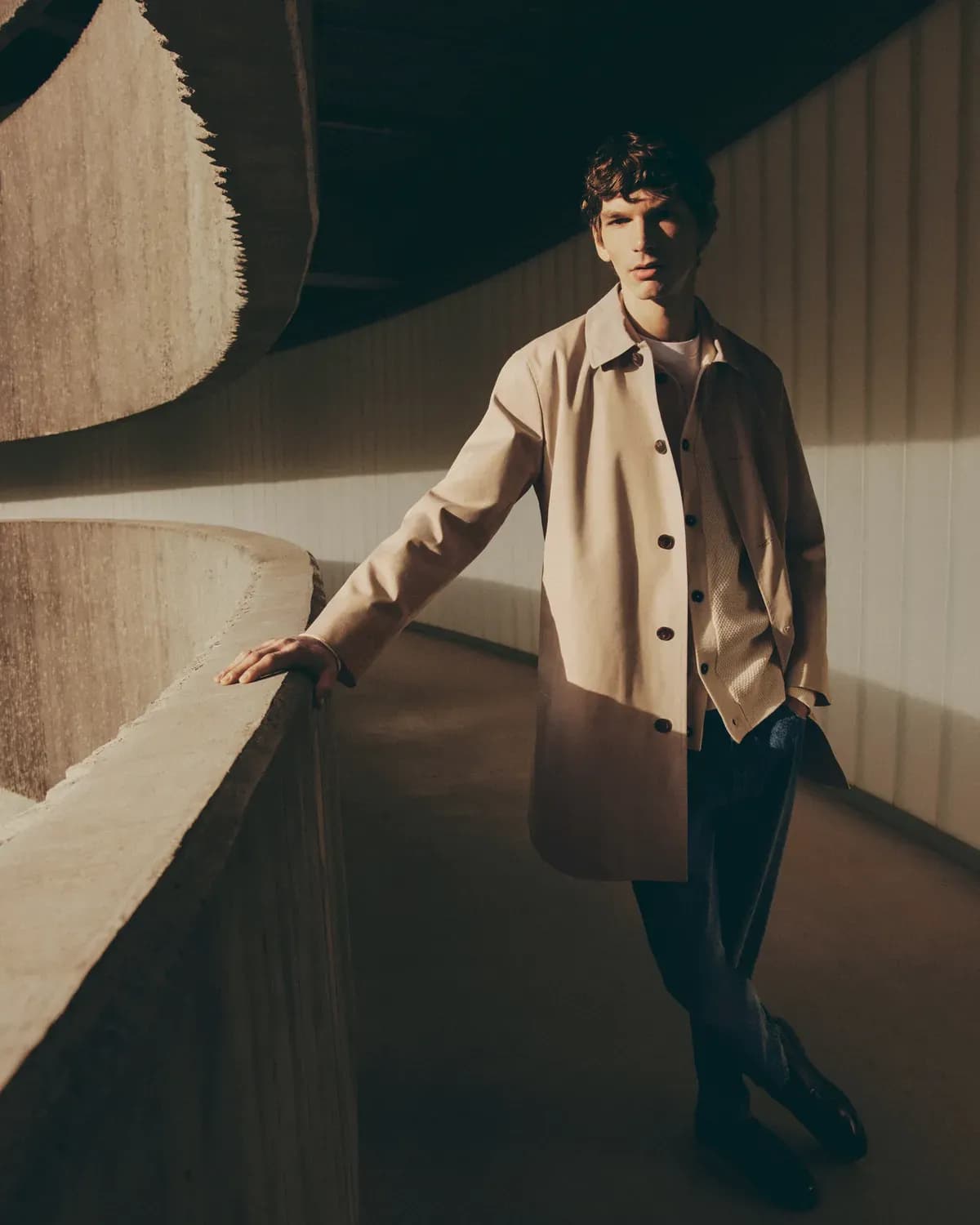 A man leaning on a curved concrete wall in a softly lit architectural space, wearing a beige coat and looking at the camera.