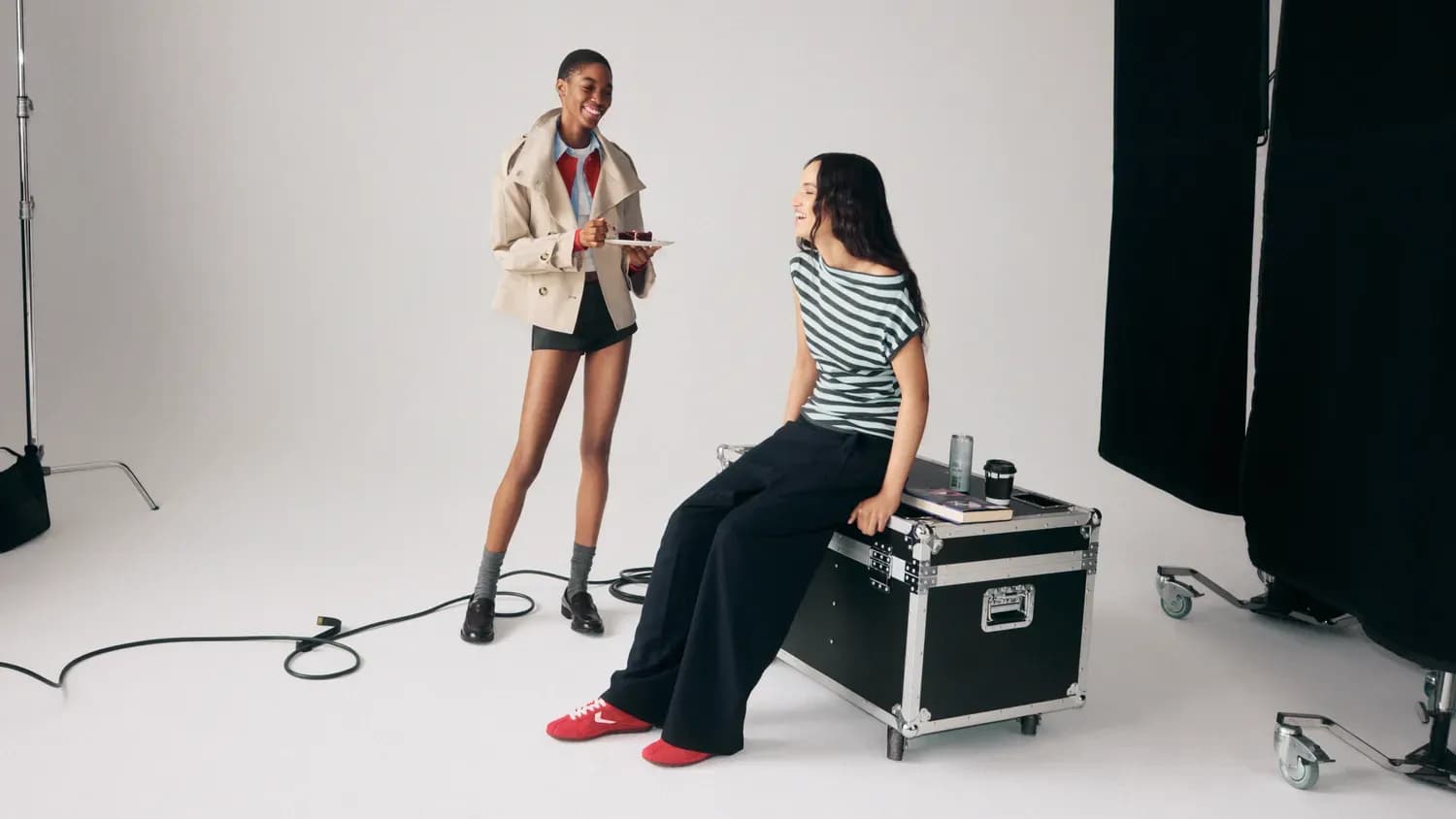 Deux femmes sourient et discutent dans un studio, l'une debout avec une assiette et l'autre assise sur une grande caisse de matériel.