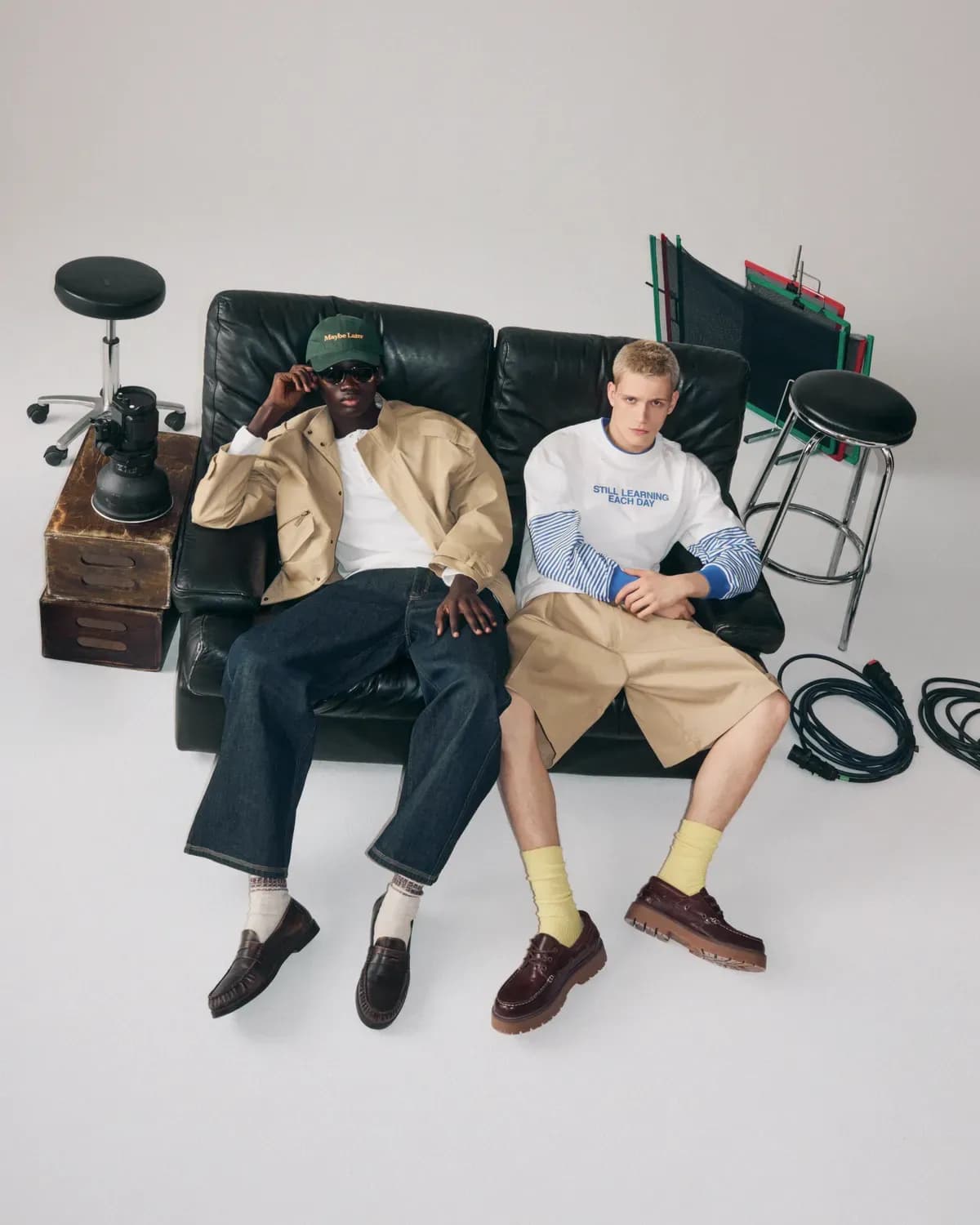 Deux hommes assis sur un canapé noir dans un studio, vêtus de tenues décontractées, entourés de matériel photographique et de câbles.