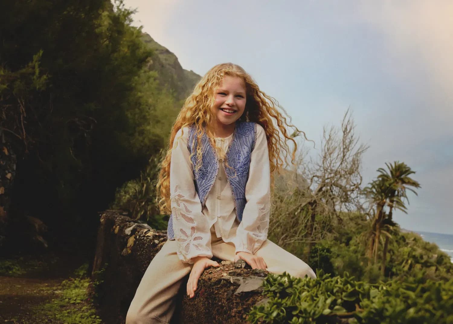 A smiling girl with long curly hair sitting on a stone wall in a lush outdoor landscape.