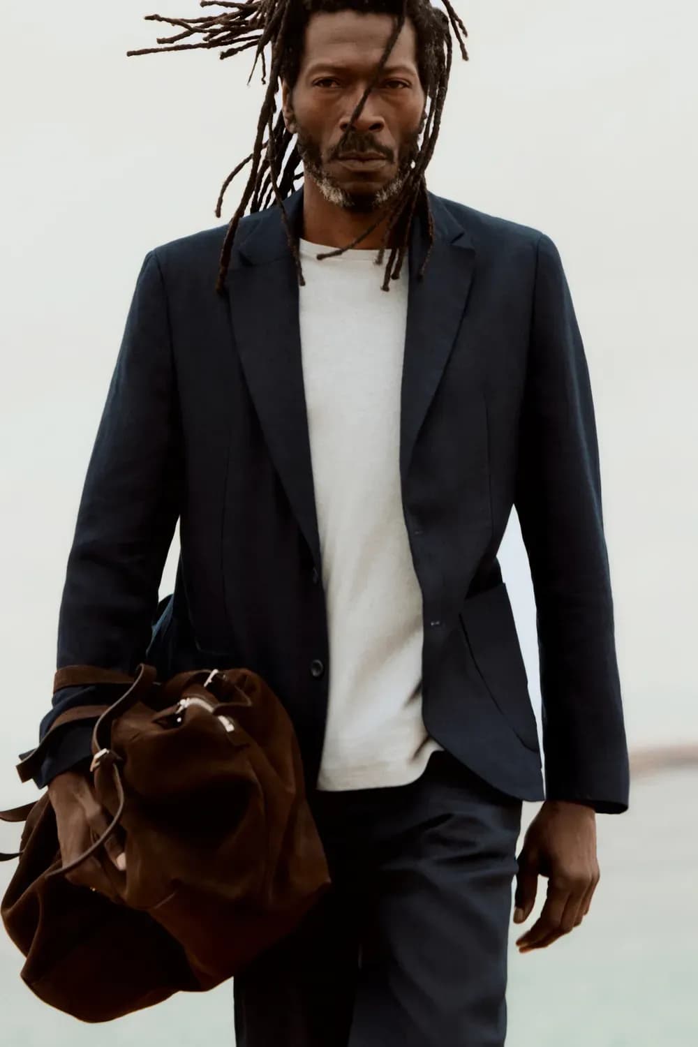 Un homme vêtu d'un costume sombre sur mesure et d'un t-shirt blanc marche à l'extérieur, portant un sac en cuir marron, ses cheveux flottant au vent sur un fond minimaliste.