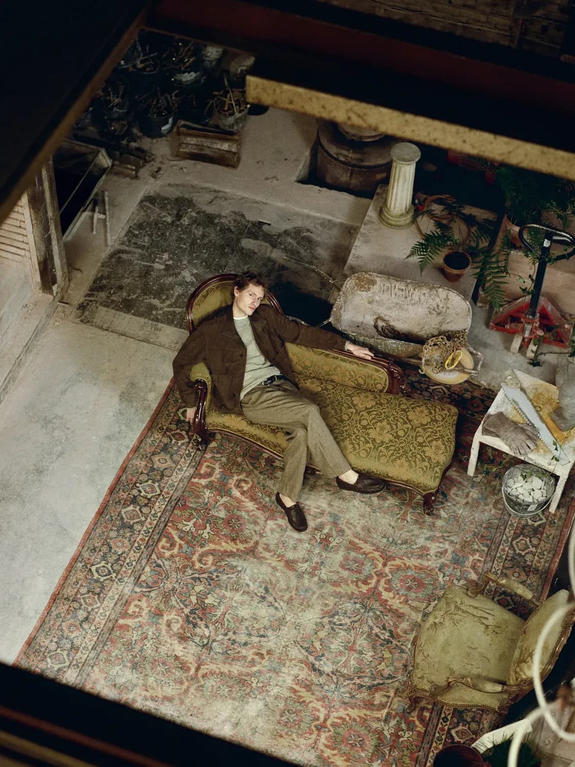 Un homme vêtu d'une veste marron, d'une chemise claire et d'un pantalon beige est allongé sur un canapé à motifs vintage dans un intérieur rustique meublé d'antiquités et agrémenté d'un grand tapis décoratif.