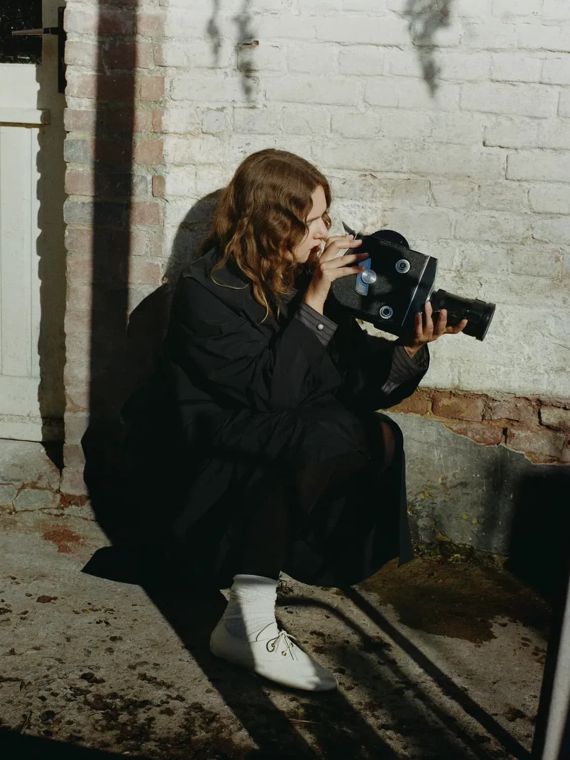Une personne vêtue d'un long manteau sombre, de chaussettes claires et de chaussures blanches est accroupie à l'extérieur, tenant un appareil photo argentique vintage, concentrée sur la prise d'une photo devant un mur texturé.