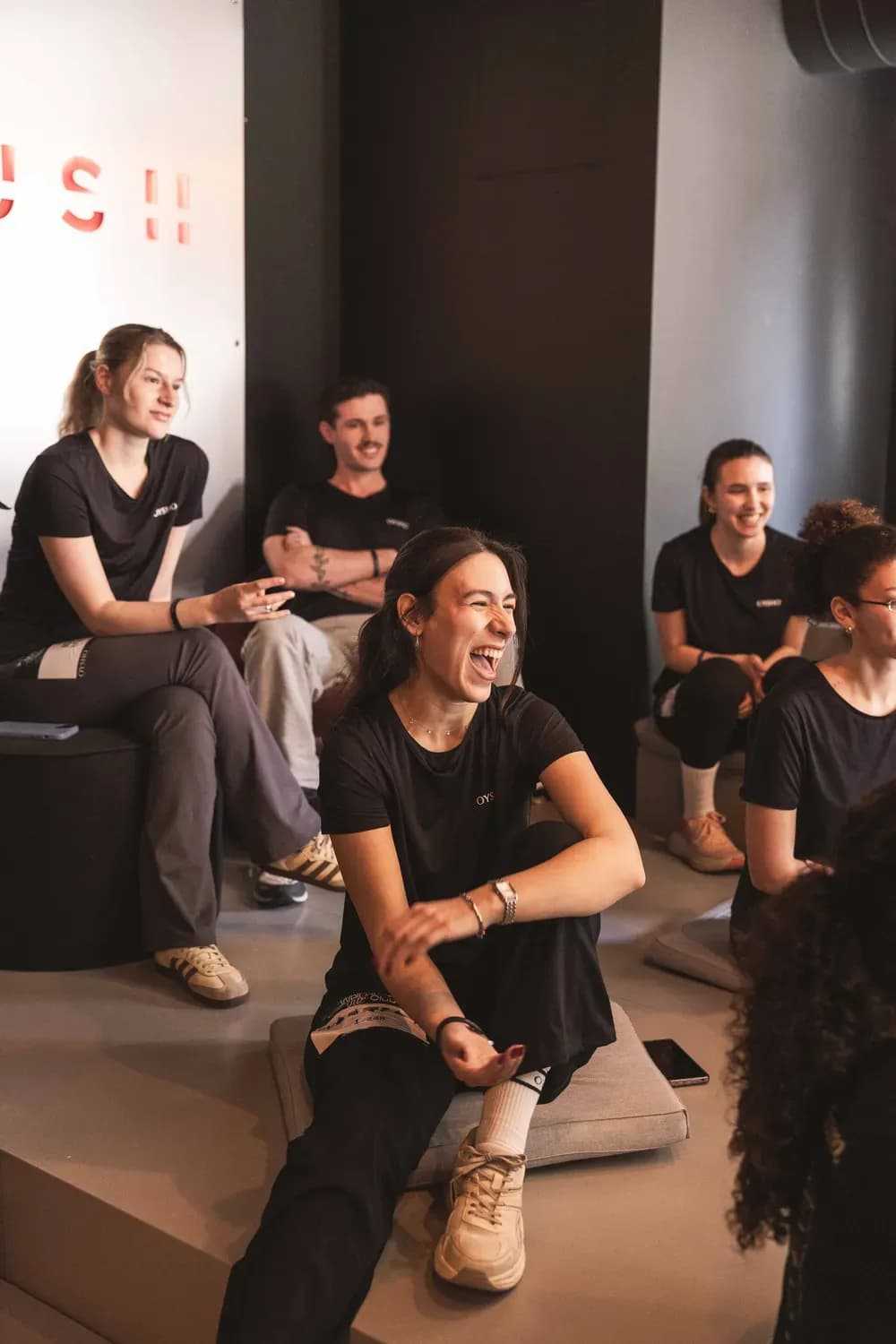 Dans un studio, un groupe de personnes vêtues de tenues de sport noires coordonnées, en t-shirts, leggings et baskets, rient ensemble dans une ambiance détendue.
