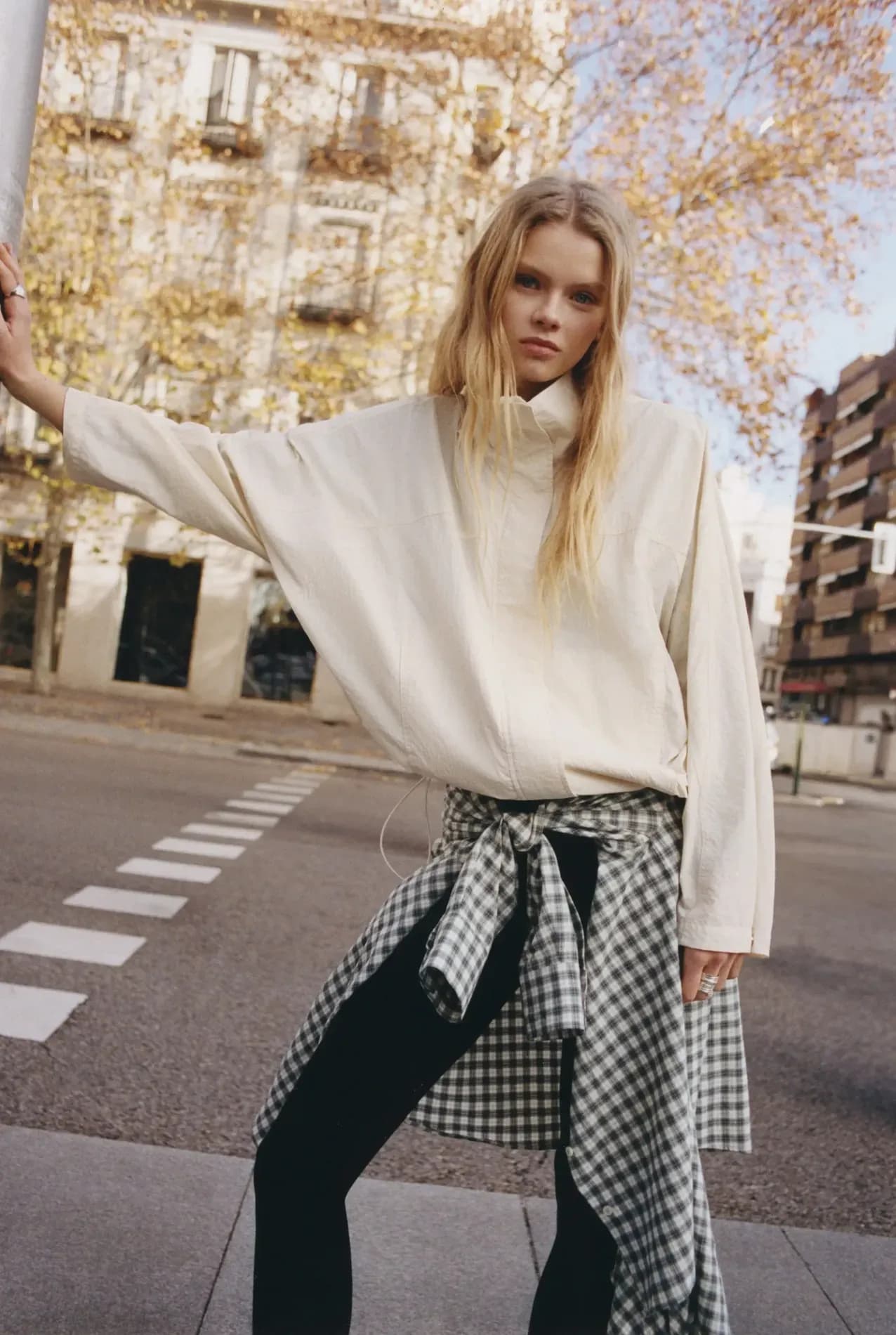 A woman wearing a loose cream jacket, black trousers, and a checkered shirt tied at the waist stands on a city street, posing in a relaxed, layered outfit.