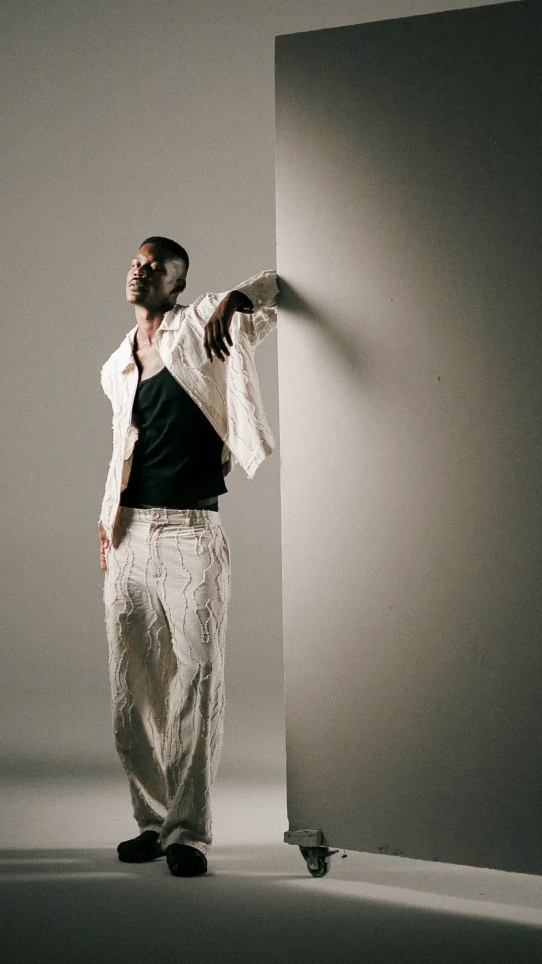 A man wears a textured light jacket and matching trousers over a dark top, creating a tonal look that emphasizes fabric detail, relaxed fit, and modern layering.