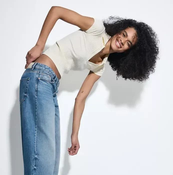 Une femme vêtue d'un haut moulant à manches courtes et d'un jean bleu pose de manière enjouée sur un fond uni.