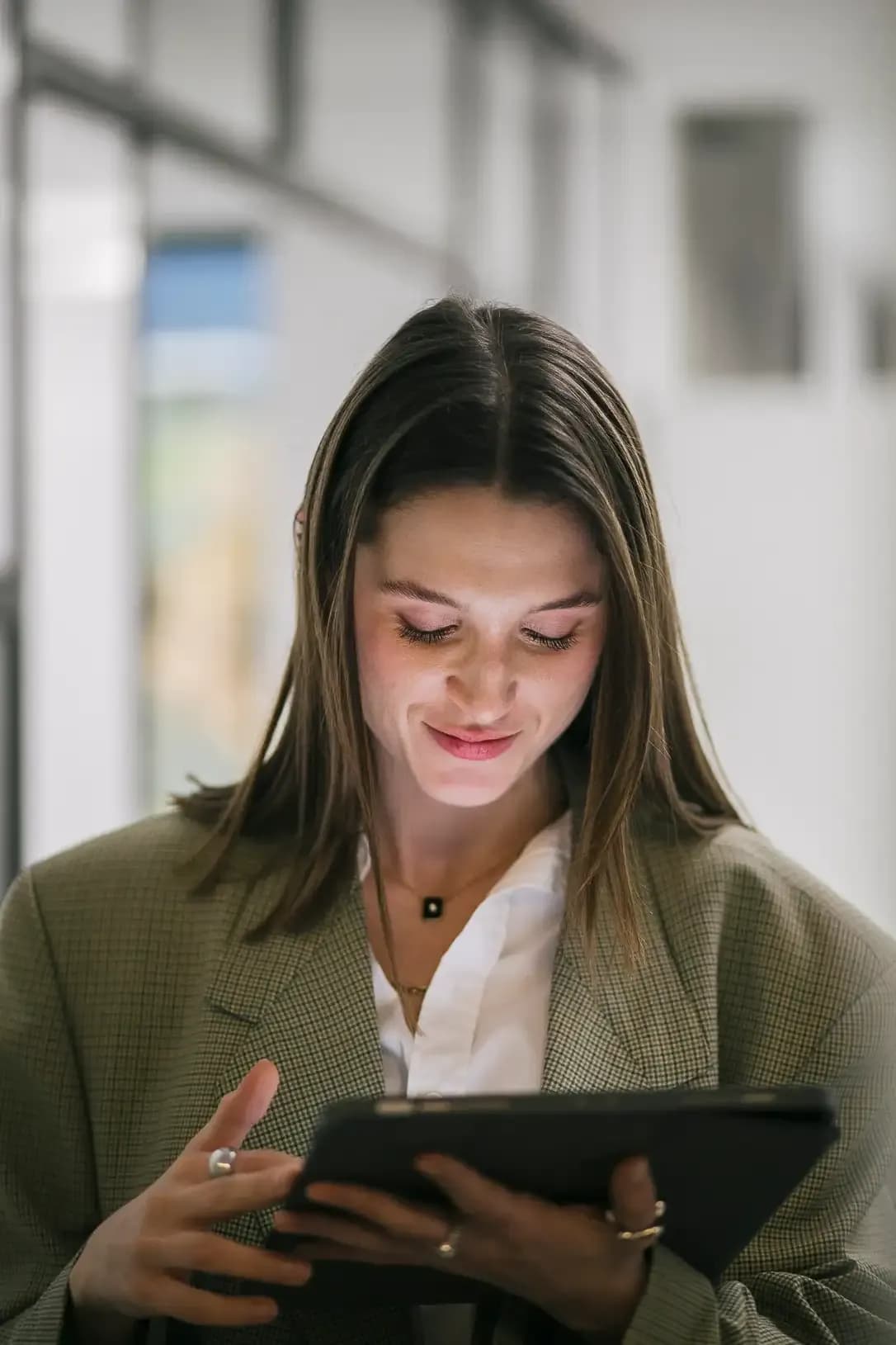 A woman wearing a tailored green blazer over a white shirt looks down at a tablet, smiling in a bright indoor setting.