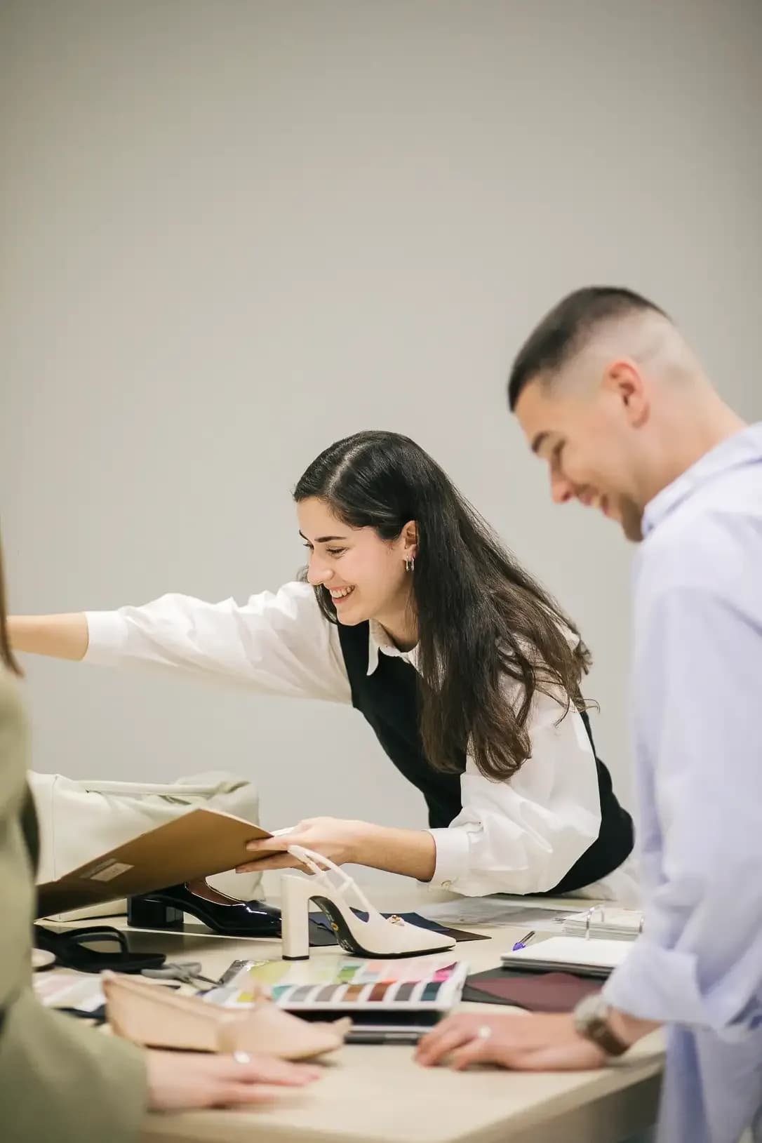 People in a fashion studio review shoe designs and materials on a table, wearing smart-casual outfits while collaborating over samples and color swatches.
