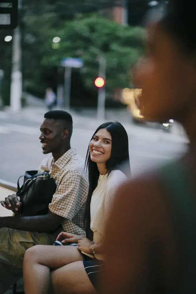 Dois amigos sentam-se ao ar livre na cidade, sorrindo e conversando enquanto esperam em uma esquina.