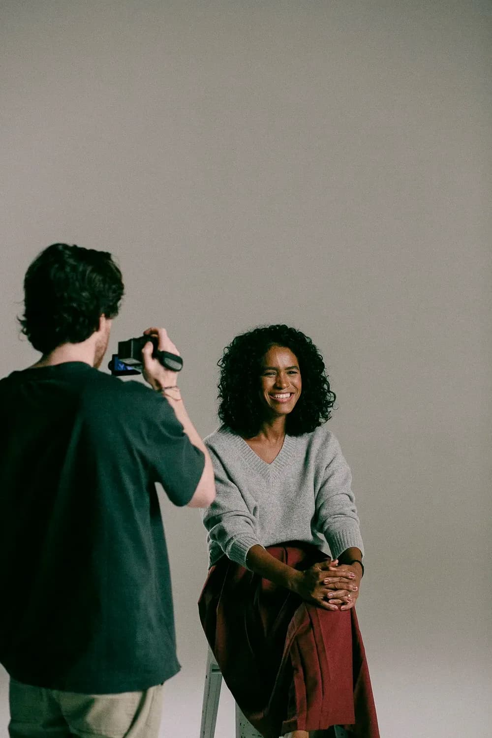Um fotógrafo filma uma mulher sorridente sentada em um banquinho contra um fundo neutro de estúdio.