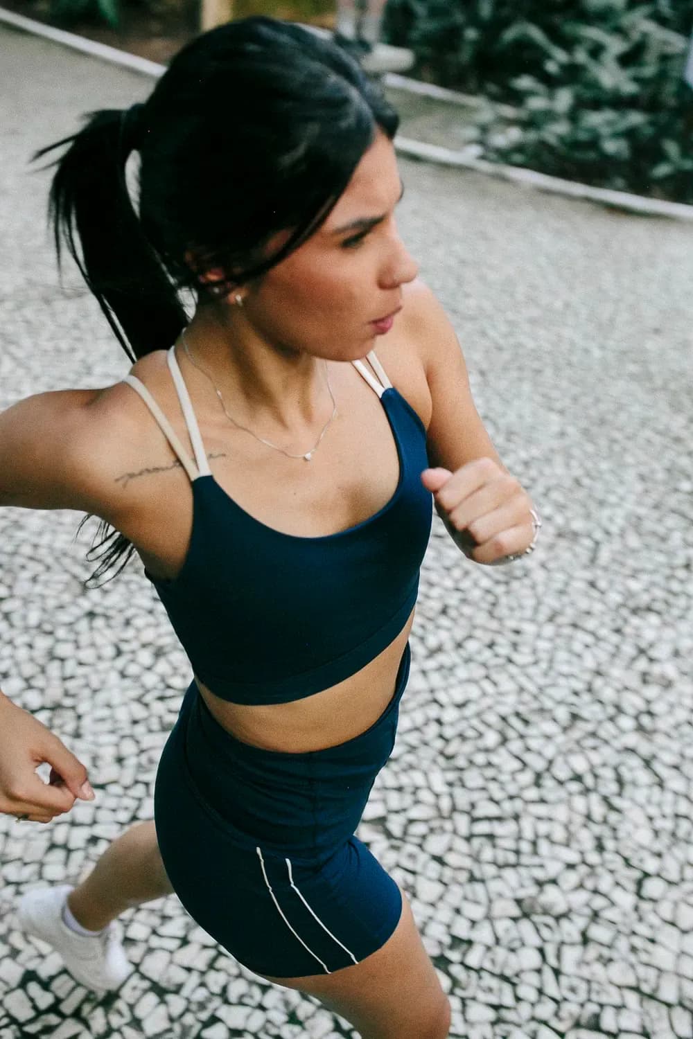Uma mulher corre ao ar livre vestindo um conjunto esportivo azul-marinho, fotografada em pleno passo em uma rua de paralelepípedos.