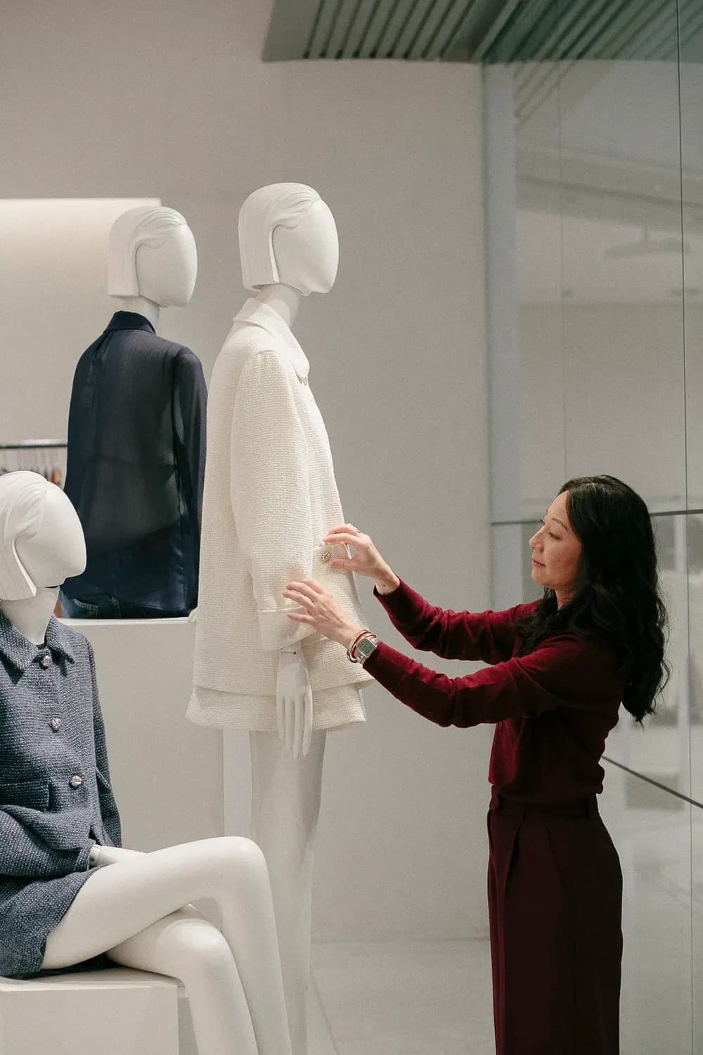 Uma estilista ajusta um conjunto creme em um manequim em um showroom moderno, cercada por outros manequins vestidos.