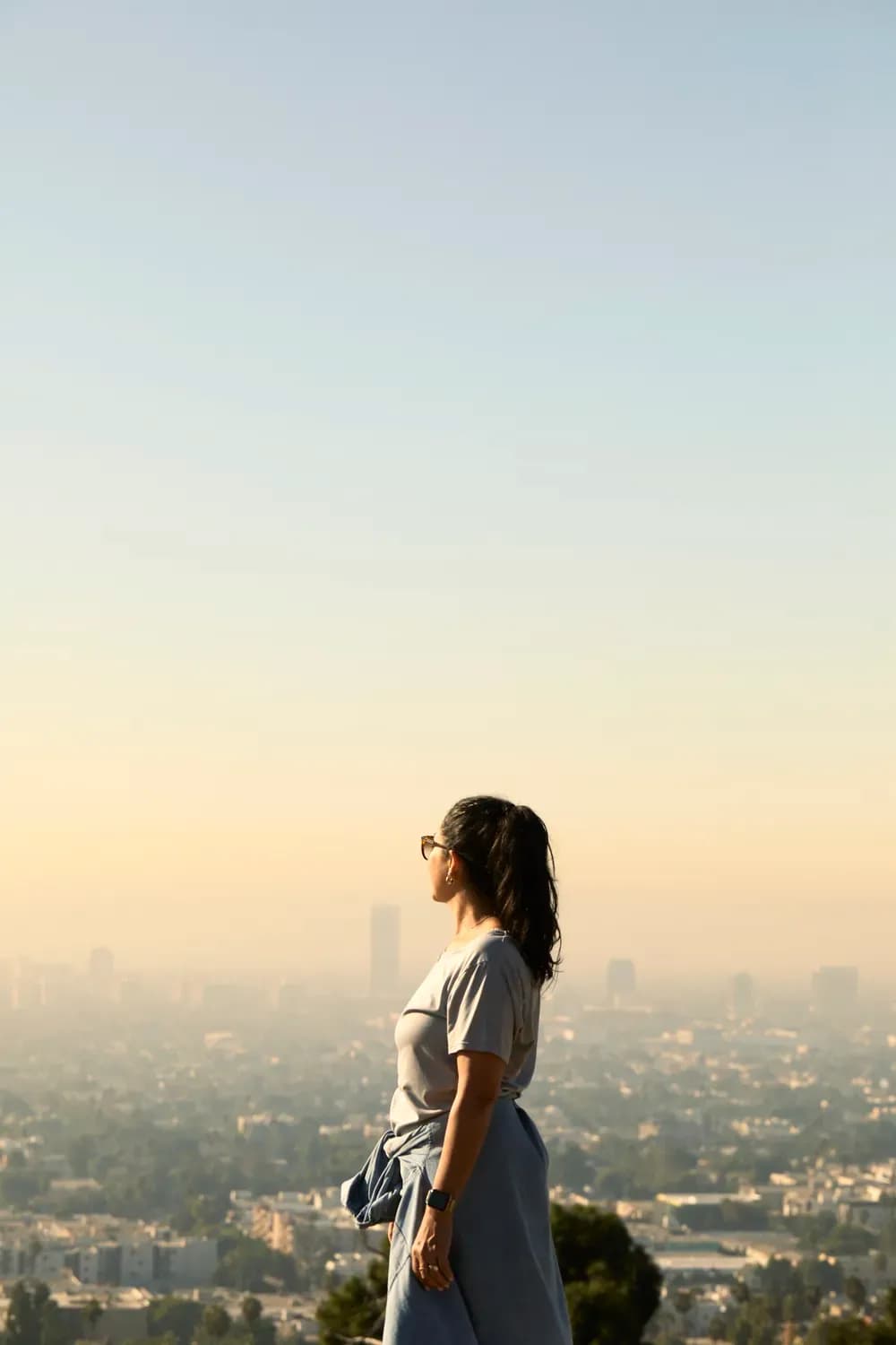 Woman in a casual t-shirt and skirt standing on a hill, overlooking a city skyline at sunset, styled in effortless everyday wear.