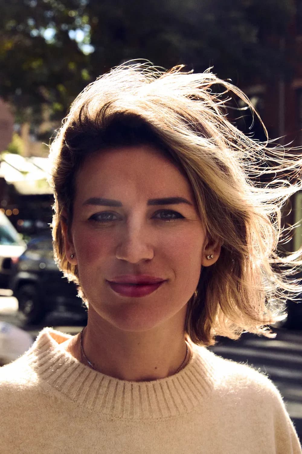 Portrait of a woman with short, windblown hair wearing a light knit sweater, standing outdoors in warm sunlight.