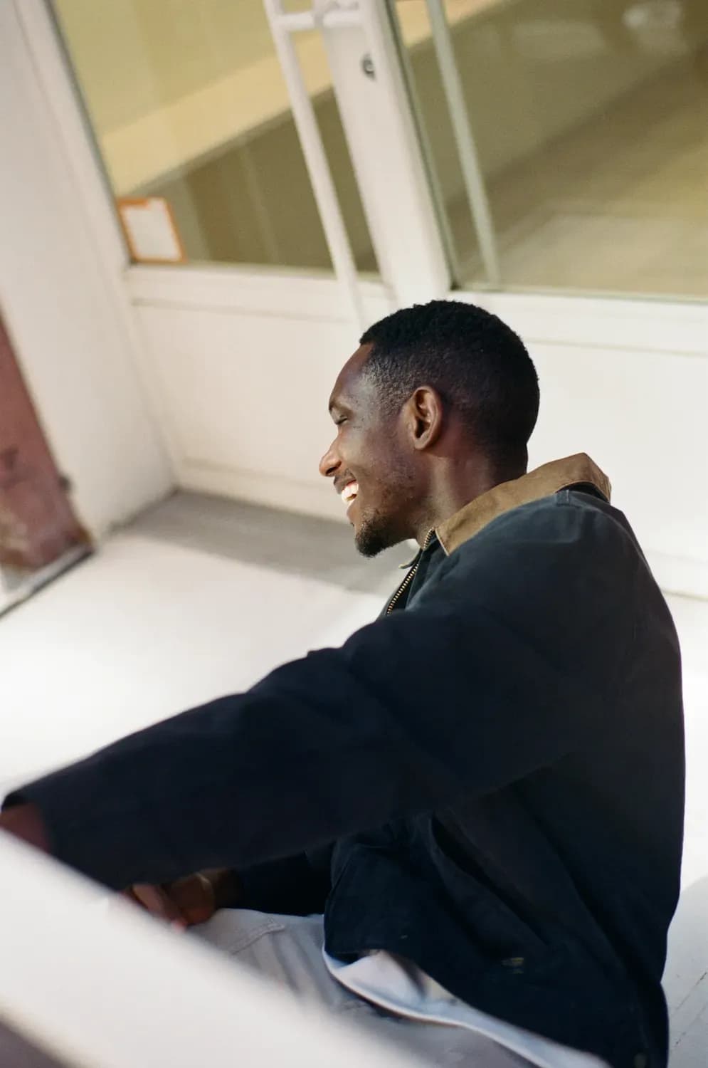 Smiling man in a dark jacket with a contrasting collar, seated indoors near large windows in a minimal setting.