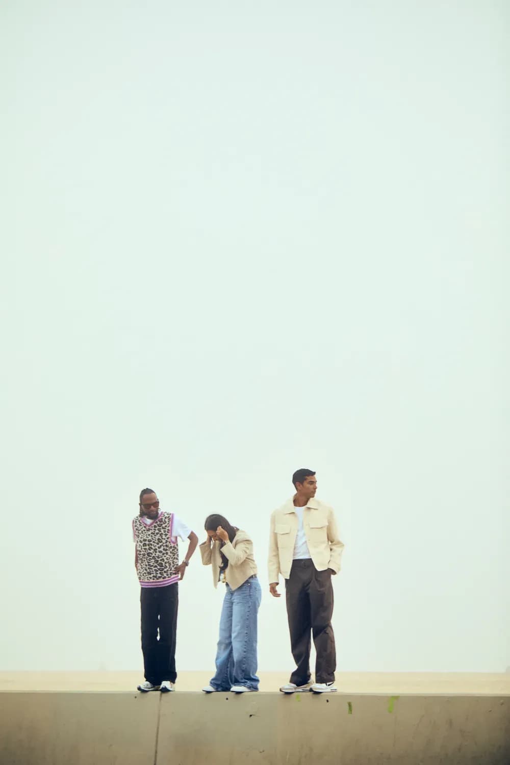 Three models standing on a ledge wearing casual, neutral-toned outfits including denim, layered tops, and a light jacket against a minimal background.