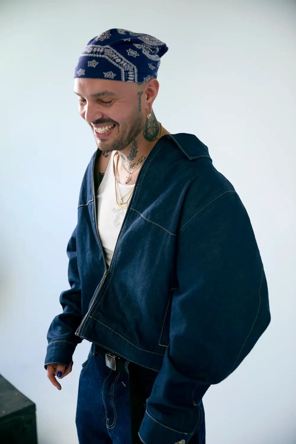 Smiling person wearing a blue patterned bandana, layered necklaces, and a dark denim jacket with matching pants against a clean background.