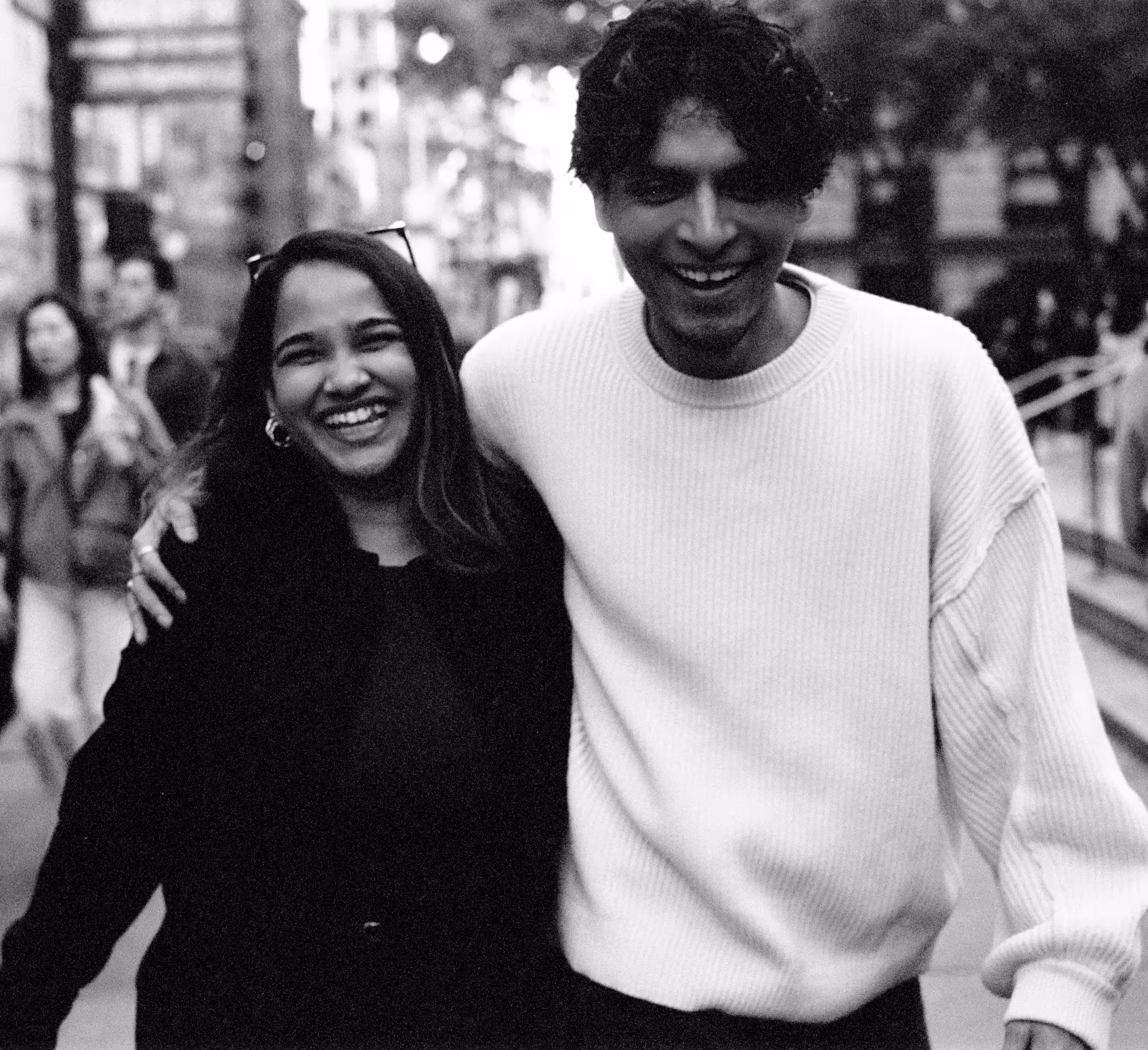 Two friends smiling and walking arm in arm on a city street, wearing casual outfits.